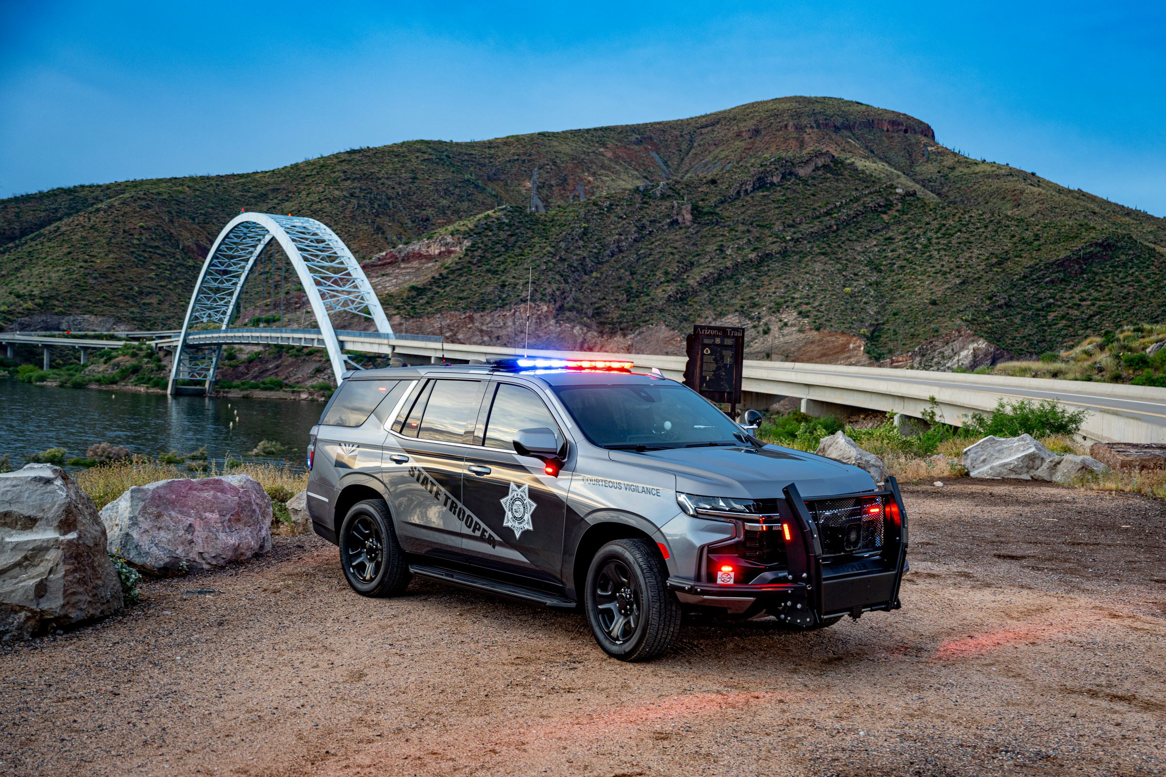 An Arizona Department of Public Safety State Trooper SUV parked near a bridge and mountains at dusk, emergency lights on